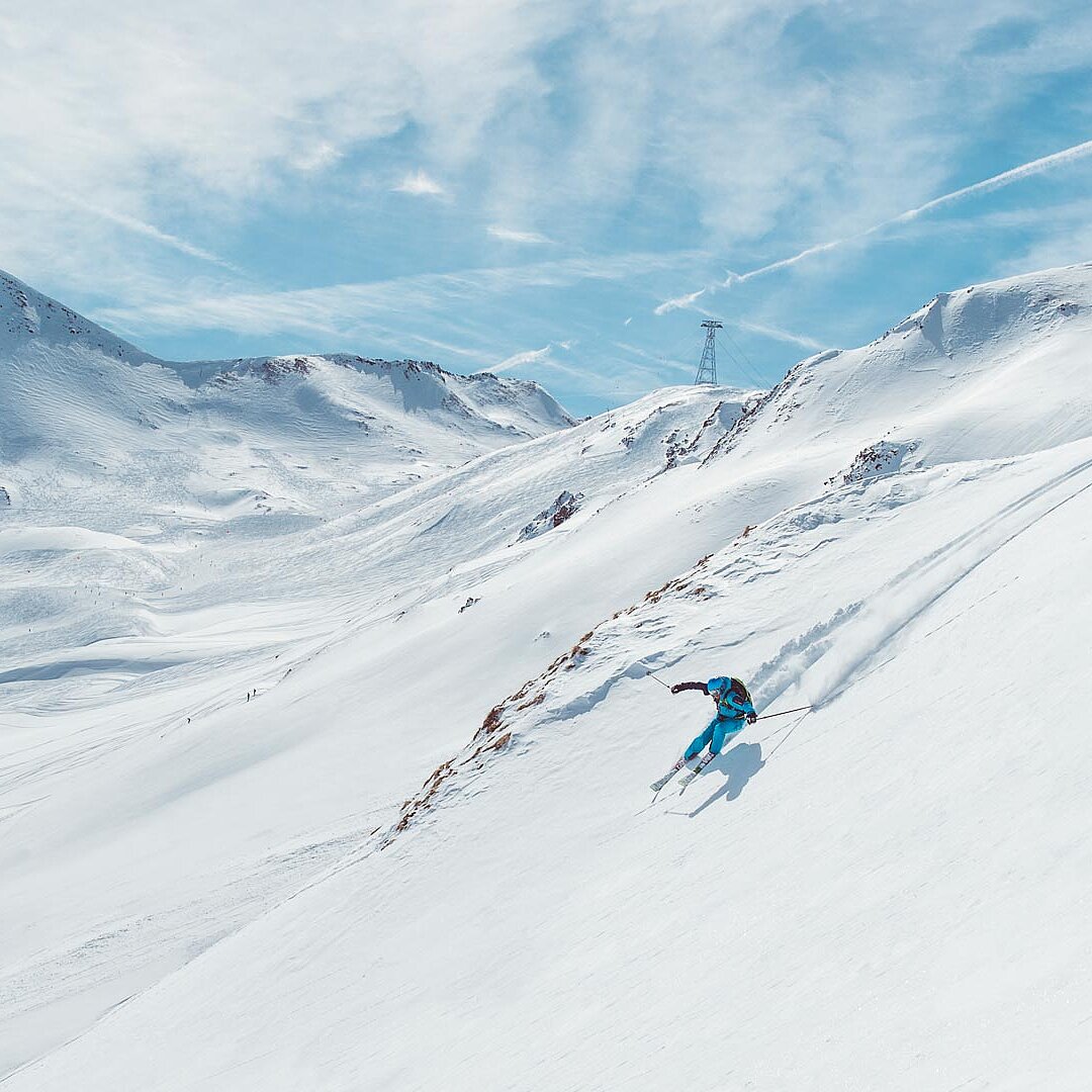 Skifahrer in türkisblau auf verschneitem Berghang in weiter Winterlandschaft mit wolkenverziertem Himmel und Alpenkulisse.