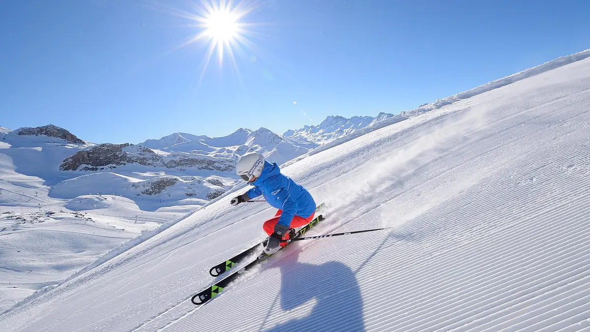 Skifahrer auf frisch präparierter Piste bei Sonnenschein, umgeben von verschneiten Bergen und klarem, blauem Himmel.